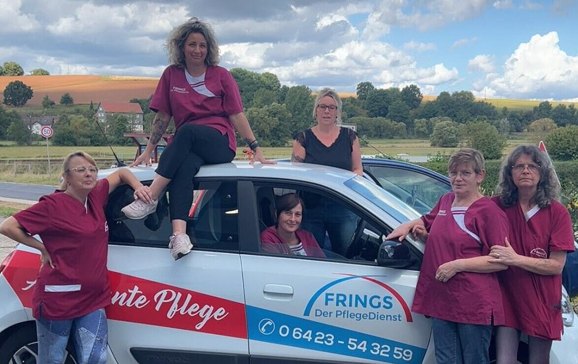Fünf Frauen in passenden kastanienbraunen Uniformen posieren in einer ländlichen Umgebung um ein kleines weißes Auto mit der Aufschrift FRINGS Der PflegeDienst.