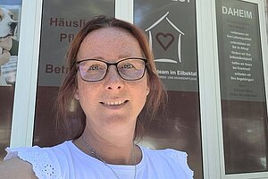 Eine Frau mit Brille und weißem Oberteil steht vor Schaufenstern, auf denen Schilder mit deutschem Text über häusliche Pflegedienste angebracht sind.