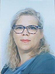 Eine Frau mit gewelltem blondem Haar, die eine Brille und ein blaues Oberteil trägt, blickt in die Kamera vor einem schlichten hellen Hintergrund.