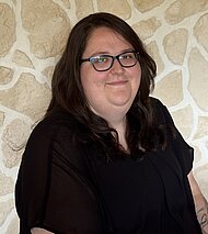 Eine Person mit langen braunen Haaren und Brille, die ein schwarzes Oberteil trägt, sitzt vor einer mit Steinen gemusterten Wand.