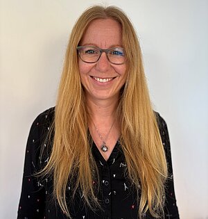 Eine Frau mit langen blonden Haaren und Brille, die ein schwarzes Oberteil mit einer Halskette trägt, steht lächelnd vor einem schlichten weißen Hintergrund.