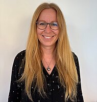 Eine Frau mit langen blonden Haaren und Brille, die ein schwarzes Oberteil mit einer Halskette trägt, steht lächelnd vor einem schlichten weißen Hintergrund.