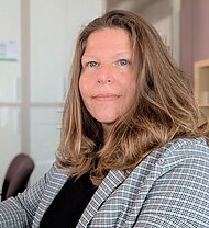 Eine Frau mit langen braunen Haaren, die einen karierten Blazer und ein schwarzes Oberteil trägt, sitzt in einem Büro und schaut in die Kamera.