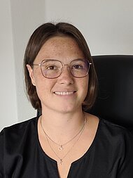 Eine Person mit kurzen braunen Haaren und Brille, die ein schwarzes Oberteil und mehrlagige Ketten trägt, sitzt auf einem Stuhl und lächelt in die Kamera.