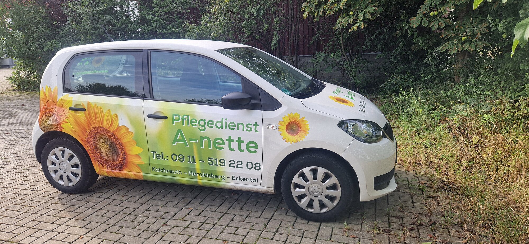 Ein weißes Auto mit einer Sonnenblumengrafik und einem Werbetext für den Pflegedienst Annette ist auf einer gepflasterten Fläche neben einer Grünanlage geparkt.