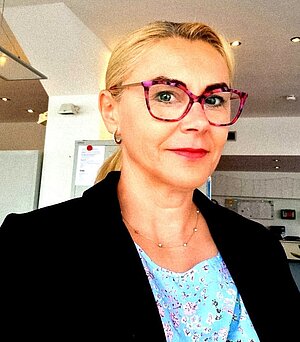 Eine Frau mit blondem Haar in einem Pferdeschwanz, roter Brille, hellblauem Oberteil und schwarzem Blazer steht mit neutraler Miene in einem Raum.