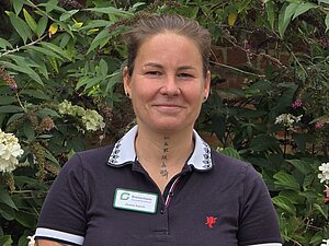 Eine Person mit hellbraunem, zurückgegeltem Haar, dunklem Kragenhemd und Namensschild steht vor grünem Laub und weißen Blumen.