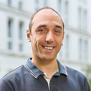 Mann mit kurzen Haaren und Geheimratsecken, dunkles Poloshirt, steht im Freien vor einem unscharfen Gebäude.