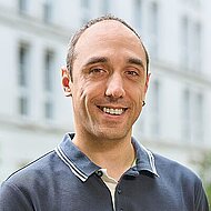 Mann mit kurzen Haaren und Geheimratsecken, dunkles Poloshirt, steht im Freien vor einem unscharfen Gebäude.