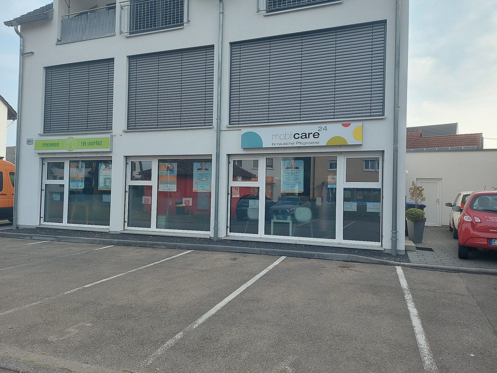 Ein zweistöckiges Gebäude mit großen Fenstern und Geschäftsschildern; davor befindet sich ein leerer Parkplatz mit markierten Stellplätzen.