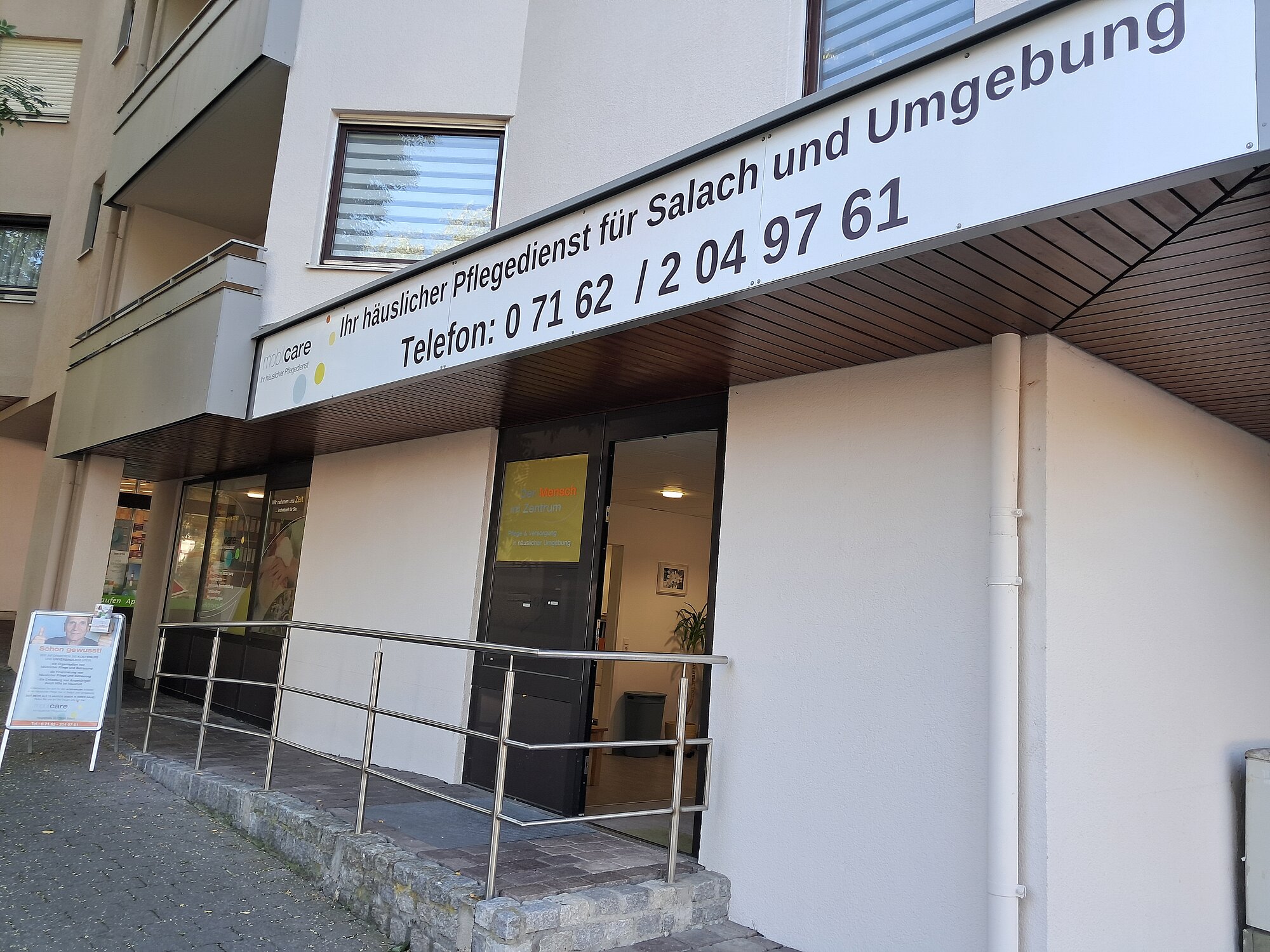 Eingang zu einem Pflegedienstbüro für Süßlach und Umgebung, mit einer Rampe, einer Glastür und einem Schild mit Kontaktinformationen über dem Eingang.