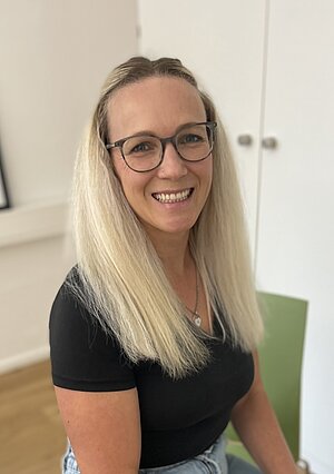 Eine Frau mit langen blonden Haaren und Brille sitzt auf einem grünen Stuhl und lächelt in einem hell erleuchteten Raum in die Kamera.