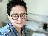 Person mit kurzen dunklen Haaren und Brille, die eine Jeansjacke trägt, steht in einem Haus vor einem Schreibtisch mit Papieren und Büroartikeln.