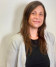 Jenny Graf mit langen braunen Haaren, einem schwarzen Oberteil und einer hellgrauen Strickjacke steht vor einer schlichten weißen Wand und lächelt leicht in die Kamera.