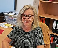 Eine Frau mit blondem Haar und Brille sitzt lächelnd an einem Bürotisch. Um sie herum befinden sich Ordner, Papiere und Büromaterial.