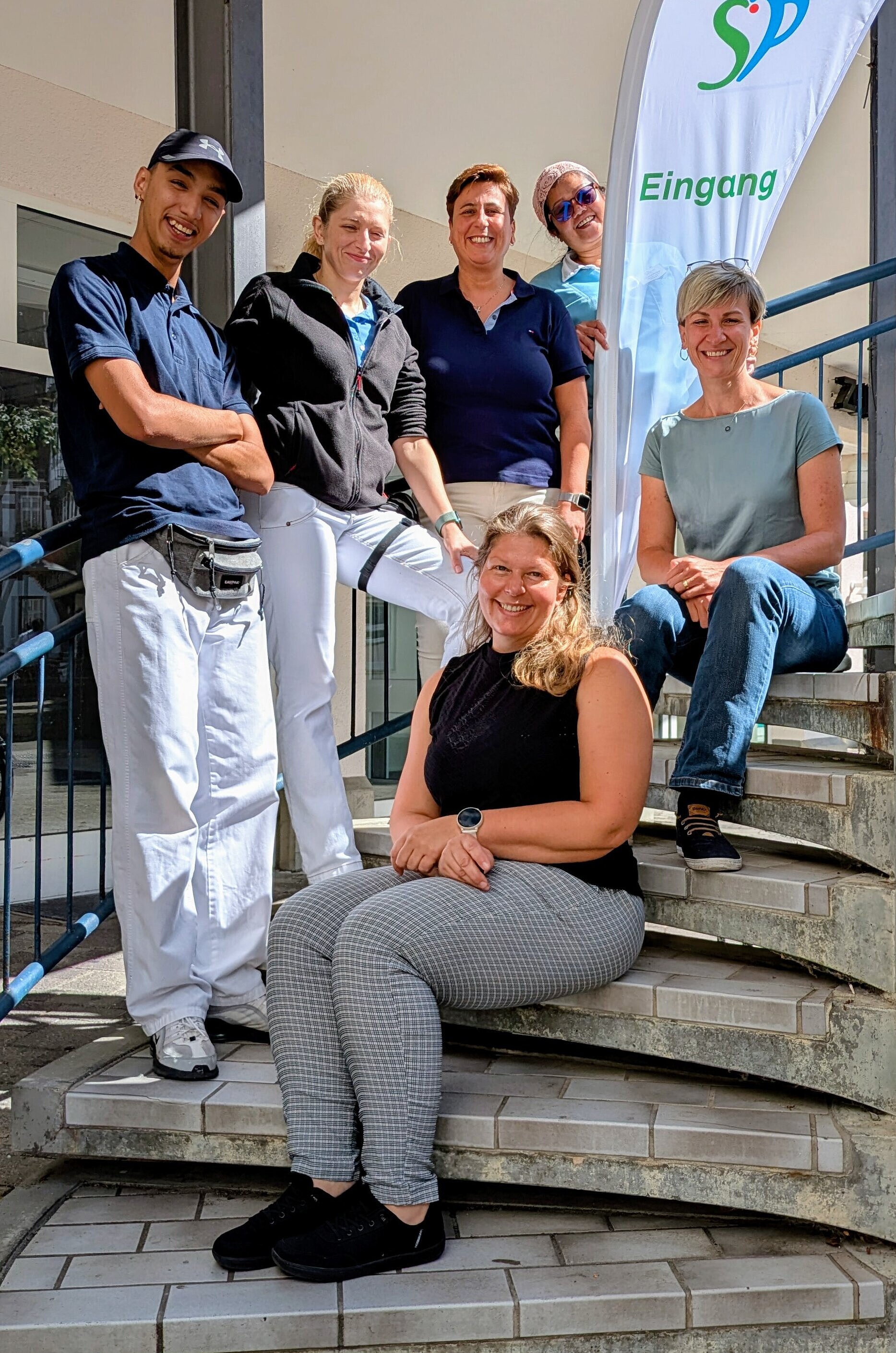 Sechs Erwachsene posieren für ein Gruppenfoto auf einer geschwungenen Treppe in einem Innenraum, mit einem Werbebanner im Hintergrund.