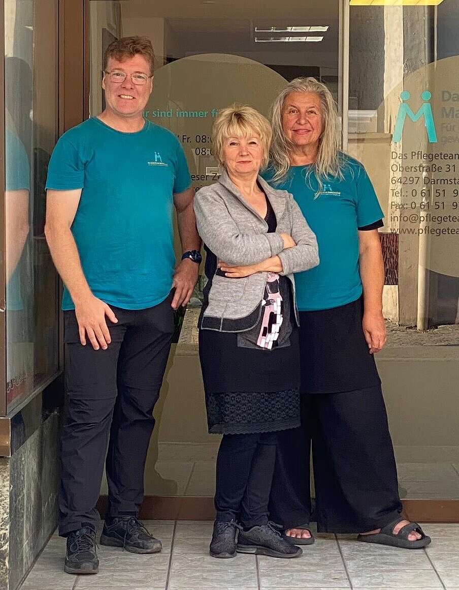 Drei Personen stehen gemeinsam vor einem gläsernen Eingang; zwei von ihnen tragen passende blaugrüne Hemden und schwarze Hosen, die dritte einen schwarzen Rock mit einer grauen Jacke.