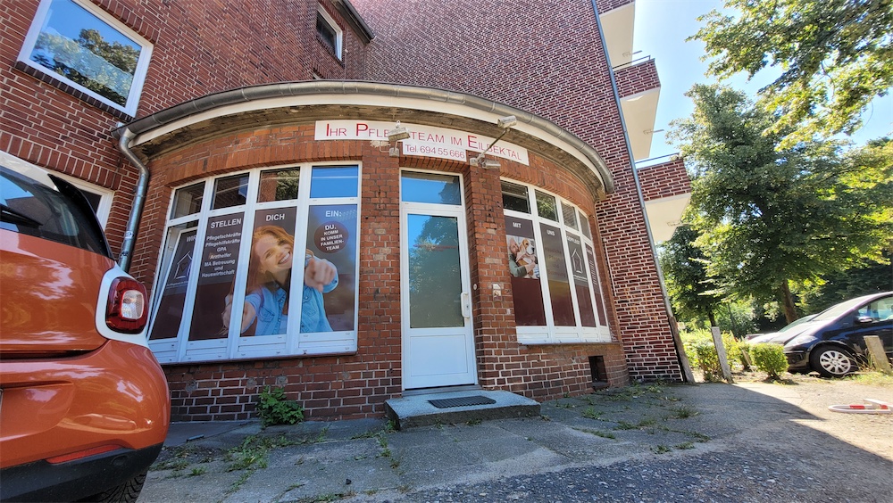 Backsteingebäude mit einem geschwungenen Eingang, großen Fenstern mit Bildern lächelnder Menschen und einer weißen Tür; in der Nähe ist ein orangefarbenes Auto geparkt, und im Hintergrund sind Bäume zu sehen.