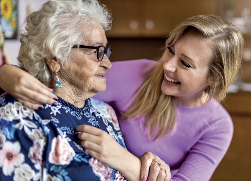 Eine ältere Frau und eine jüngere Frau sitzen eng beieinander, lächeln und teilen einen fröhlichen Moment in einem Haus.