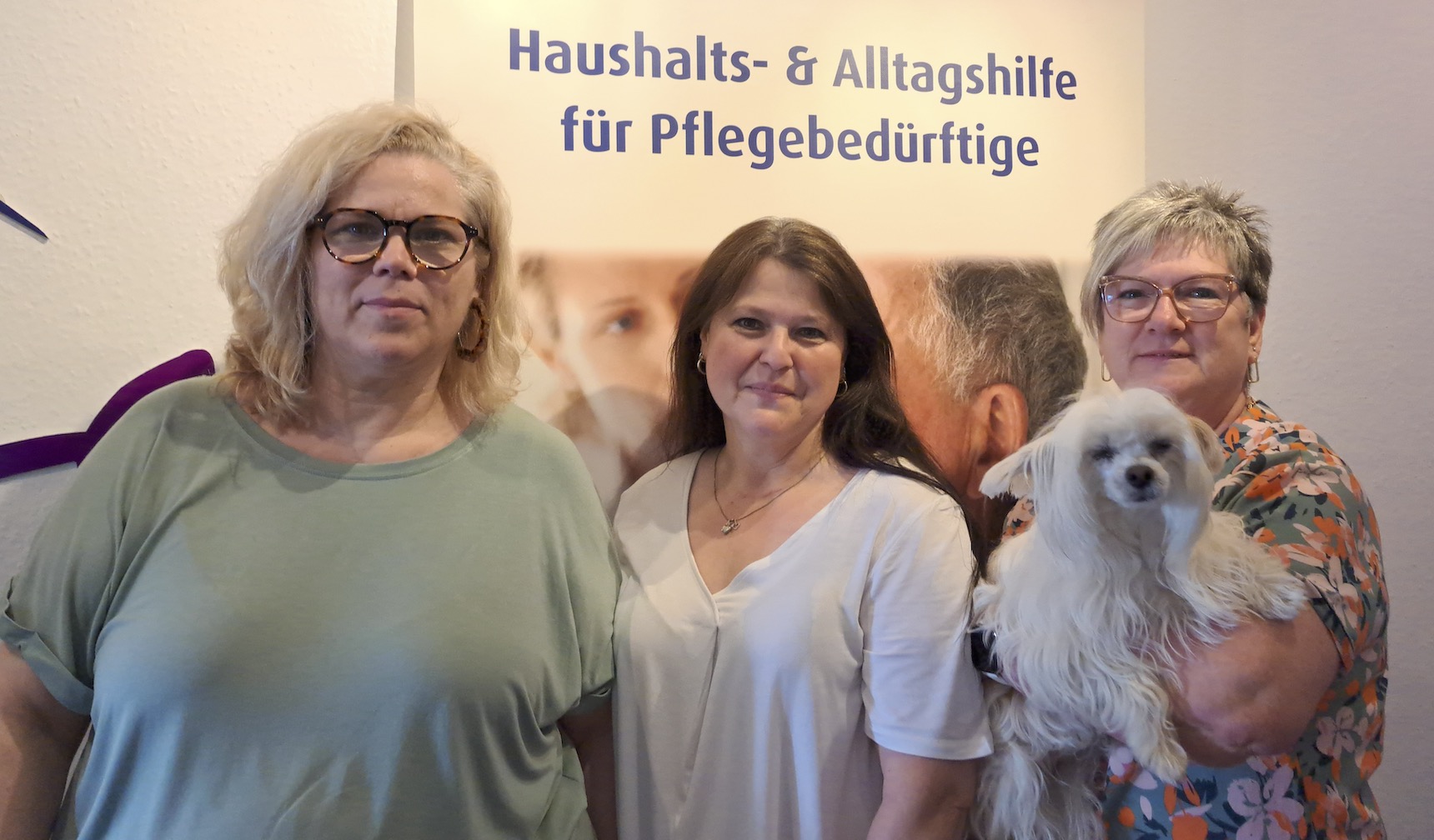 Drei Frauen stehen vor einem Schild über Haushalts- und Alltagshilfen für Pflegebedürftige; eine Frau hält einen kleinen weißen Hund.